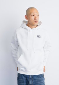 Hoodie branco feito de um tecido suave, com um bolso frontal, apresentando um pequeno logo da Nike em azul marinho no lado esquerdo do peito. Usado com jeans azuis.