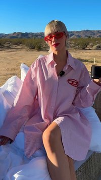 Persoană cu părul scurt blond și ochelari de soare roșii, așezată în aer liber, îmbrăcată într-o rochie în dungi roz, ținând o cană neagră în fața unui peisaj de deșert.