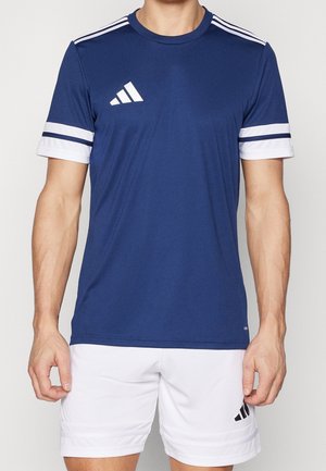 Homme portant un maillot de sport bleu marine avec des rayures blanches sur les épaules et les manches, assorti à un short blanc arborant un logo noir.