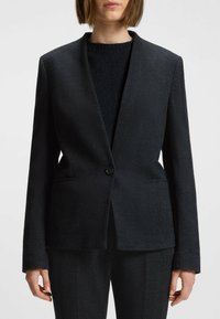 Sort herringbone blazer med en enkelt knaplukning, klapkrave, to frontlommer og en skræddersyet silhuet. Bæres over en mørk striktrøje.
