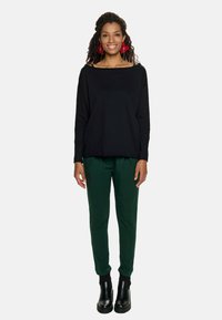 Czarny top z długim rękawem i fasonem typu off-shoulder, połączony z zielonymi wąskimi spodniami. Czarne botki za kostkę i czerwone kwieciste kolczyki dopełniają stylizację.