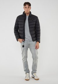 Giovane uomo che indossa un giubbotto a vento nero, una maglietta grigia, jeans grigi chiari e sneakers bianche, in piedi con una mano in tasca contro uno sfondo bianco.