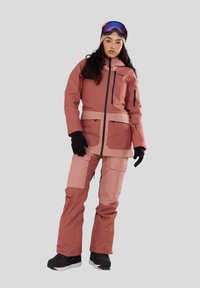 Roze en lichtroze winterjack met een capuchon, rits aan de voorkant en zakken. Gecombineerd met bijpassende roze skibroek en zwarte handschoenen.