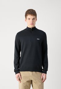 Sudadera negra con cuello de media cremallera, textura acanalada en los hombros y un logo en el lado izquierdo del pecho. Tejido suave, mangas largas y bajo entallado.