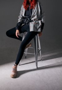 Rutig blazer över svart sweatshirt, kombinerat med svarta leggings och klumpiga rosa sneakers. Modellen sitter på en metallstol.