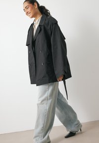 Czarny dwurzędowy płaszcz trench z naramiennikami, dużymi guzikami i przednimi kieszeniami, w połączeniu z jasnoniebieskimi spodniami typu wide-leg.