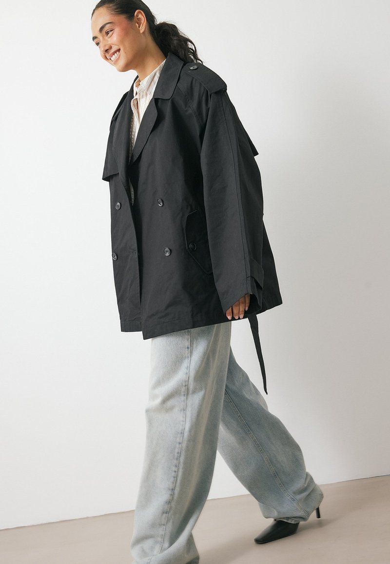 Czarny dwurzędowy płaszcz trench z naramiennikami, dużymi guzikami i przednimi kieszeniami, w połączeniu z jasnoniebieskimi spodniami typu wide-leg.