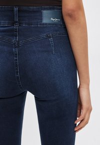 Vaqueros de mezclilla azul oscuro con un diseño ajustado, que presentan una etiqueta de cuero en la cintura y bolsillos traseros estándar. Textura suave.