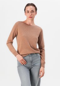 Brązowy sweter wykonany z miękkiego materiału, z luźnym dekoltem i długimi rękawami, połączony z jasnoszarymi dżinsami.