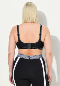 Sostenedor deportivo negro con tirantes ajustables y cierre de gancho y ojal. Presenta costuras grises y una textura suave, combinado con leggings negros.