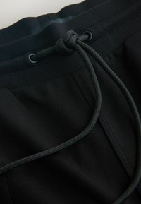 Close-up of black sweatpants waistband with drawstring tied in a knot, showing fabric texture and stitching details.