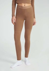 Bruine aansluitende leggings van gladde rekbare stof, met een hoge tailleband en een naadloos ontwerp. Draagt witte enkelsokken.