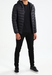 Schwarze Steppjacke mit Kapuze, gesteppter Musterung, vorderem Reißverschluss. Wird über einem schwarzen T-Shirt getragen und mit schwarzer Jeans sowie schwarzen Sneakers kombiniert.