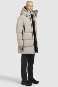 Beige puffjakke med høj krave, lynlås-lommer og en let skinnende tekstur. Båret med sorte jeans og sorte sko.
