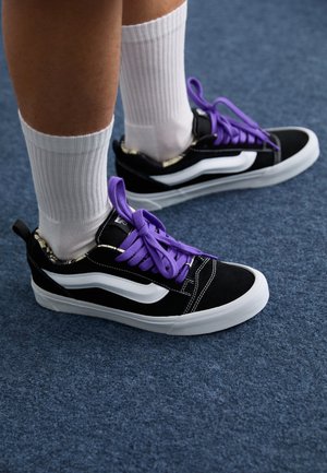 Persoon draagt zwart-witte sneakers met paarse veters en witte geribbelde crew sokken, staand op een blauw tapijt.