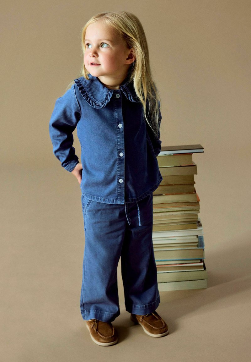 Jeune fille en chemise bleue et pantalon se tient près d'une haute pile de livres sur un fond beige uni.