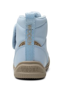 Bota azul de cuero hasta el tobillo con una suela de goma beige texturizada. Presenta una correa trasera trenzada con la marca "WODEN" y un collar suave acolchado.