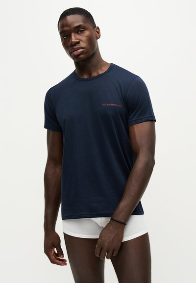 Νέος άνδρας με μπλε σκούρο t-shirt Emporio Armani και λευκά εσώρουχα boxer, στέκεται μπροστά σε ένα απλό ανοιχτό φόντο.