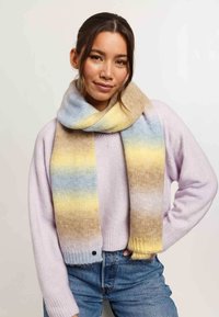 Sciarpa lavorata a maglia con una gradazione di blu, giallo e beige, caratterizzata da una consistenza morbida. Il modello indossa un maglione viola chiaro e dei jeans in denim.