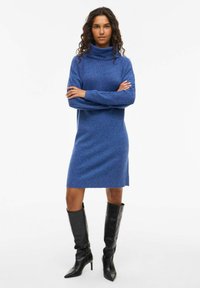 Blå stickad klänning med polokrage och lång ärm. Figursydd design som slutar ovanför knäna. Bärs med svarta knähöga klackstövlar.