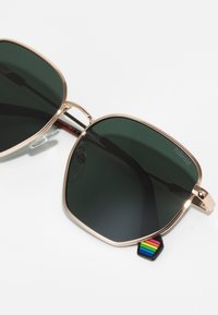 Ochelari de soare cu o ramă din metal auriu, lentile colorate în verde, formă pătrată și un etichetă în dungi multicolore atașată de braț.