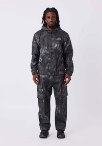 Set composto da giacca nera con effetto tie-dye e pantaloni cargo, caratterizzato da chiusura con zip e numerose tasche, realizzato in un materiale liscio e testurizzato.