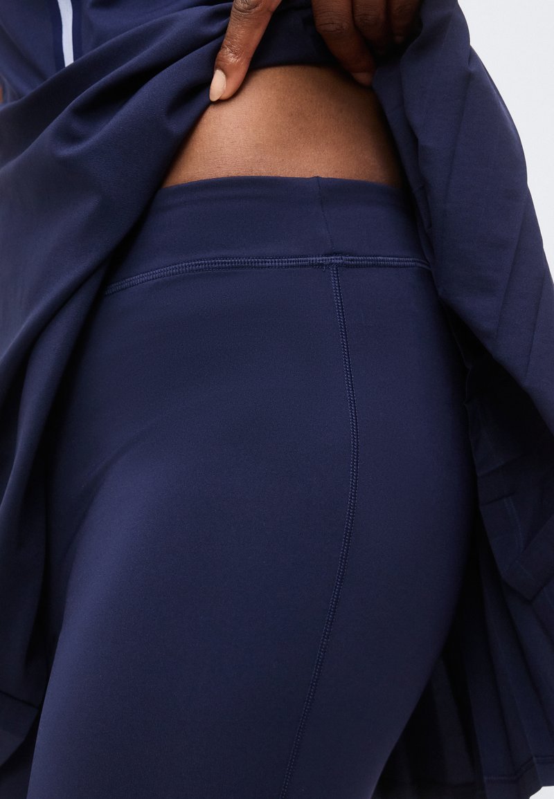 Dunkelhäutige Hand hebt marinefarbener Oberteil an, um marinefarbene Leggings mit hohem Bund und sichtbaren Nahtdetails an Hüfte und Oberschenkel zu enthüllen.