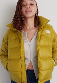 Puhasta jakna v olivno zeleni barvi, z visokim ovratnikom, zadrgo in horizontalnim šivanjem. Logotip je prikazan zgoraj levo.