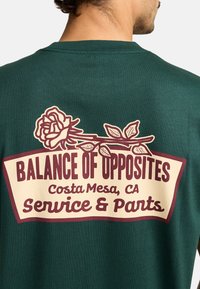 Camiseta de manga corta verde oscuro con un gráfico en crema y burdeos que presenta una ilustración de una rosa y el texto "Balance de Opuestos."