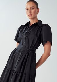 Juoda marškininė suknelė su trumpomis pūkuotomis rankovėmis, susiaučiamu liemeniu ir sluoksniuota sijonė. Lygus audinys su subtiliu blizgesiu ir be matomų raštų.