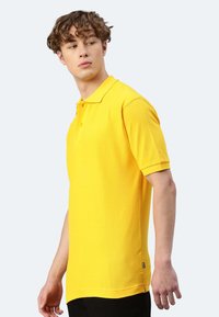 Gelbes Polohemd aus strukturiertem Stoff, mit klassischem Kragen und kurzen Ärmeln, zwei Knöpfen und einem kleinen Logohängers am Saum.