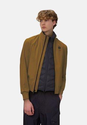 Giovane con capelli ricci che indossa una giacca color senape sopra una giacca nera trapuntata con zip, con le mani nelle tasche dei pantaloni cargo scuri, guarda di lato.