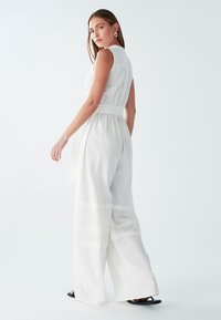 Mono blanco sin mangas con cintura ceñida, diseño de pierna ancha y tela transparente. Presenta rayas horizontales y un cuello.