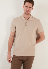 Béžové polo tričko s límečkem, zapínáním na tři knoflíky a krátkým rukávem. Vyrobeno z texturované tkaniny s jemnými švy. V kombinaci s hnědými kalhotami.