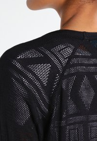 Black knitted cardigan featuring an openwork pattern with geometric shapes, showcasing a blend of solid and mesh textures on the back.