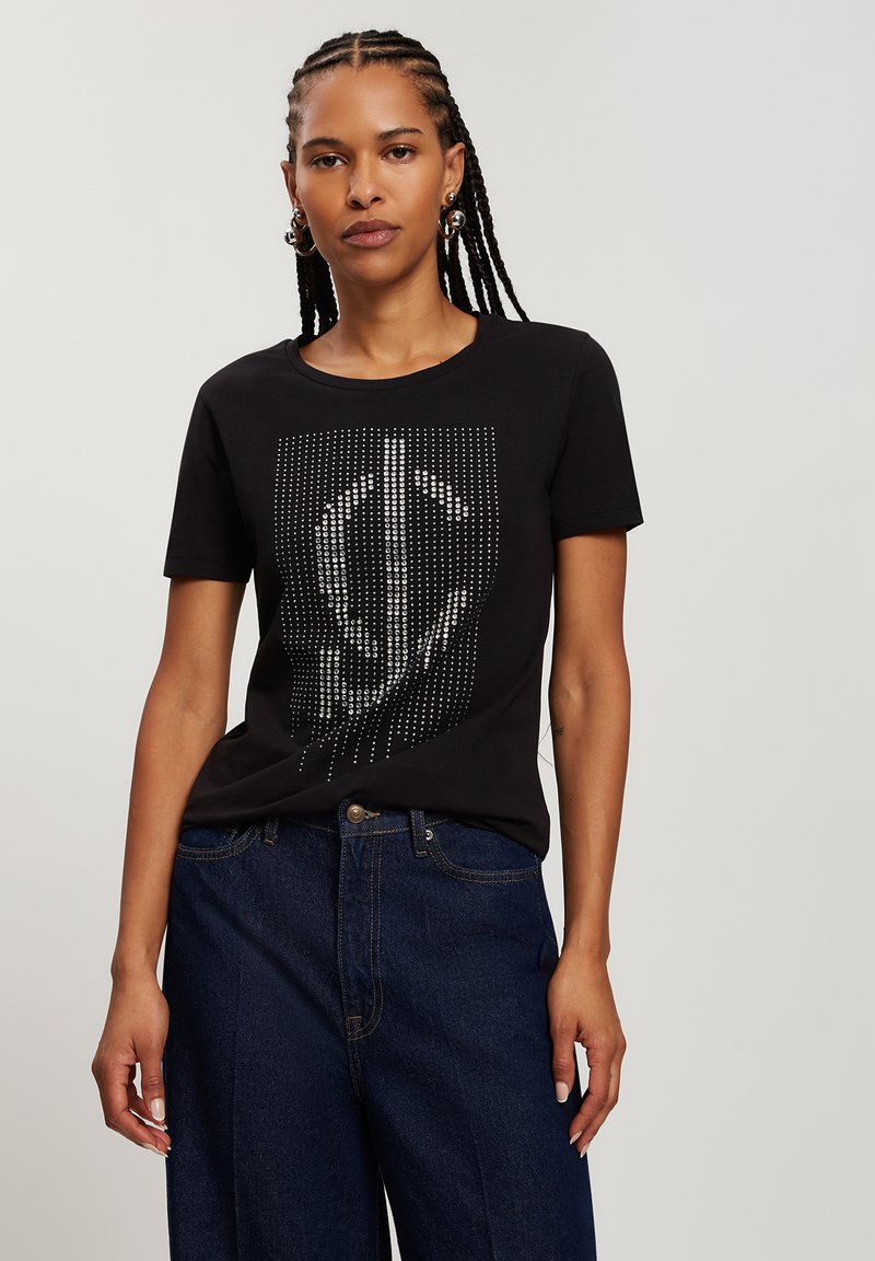 Czarna bawełniana koszulka z krótkim rękawem, ozdobiona graficznym wzorem złożonym ze srebrnych ćwieków, z okrągłym dekoltem. Noszona z luźnymi niebieskimi dżinsami.