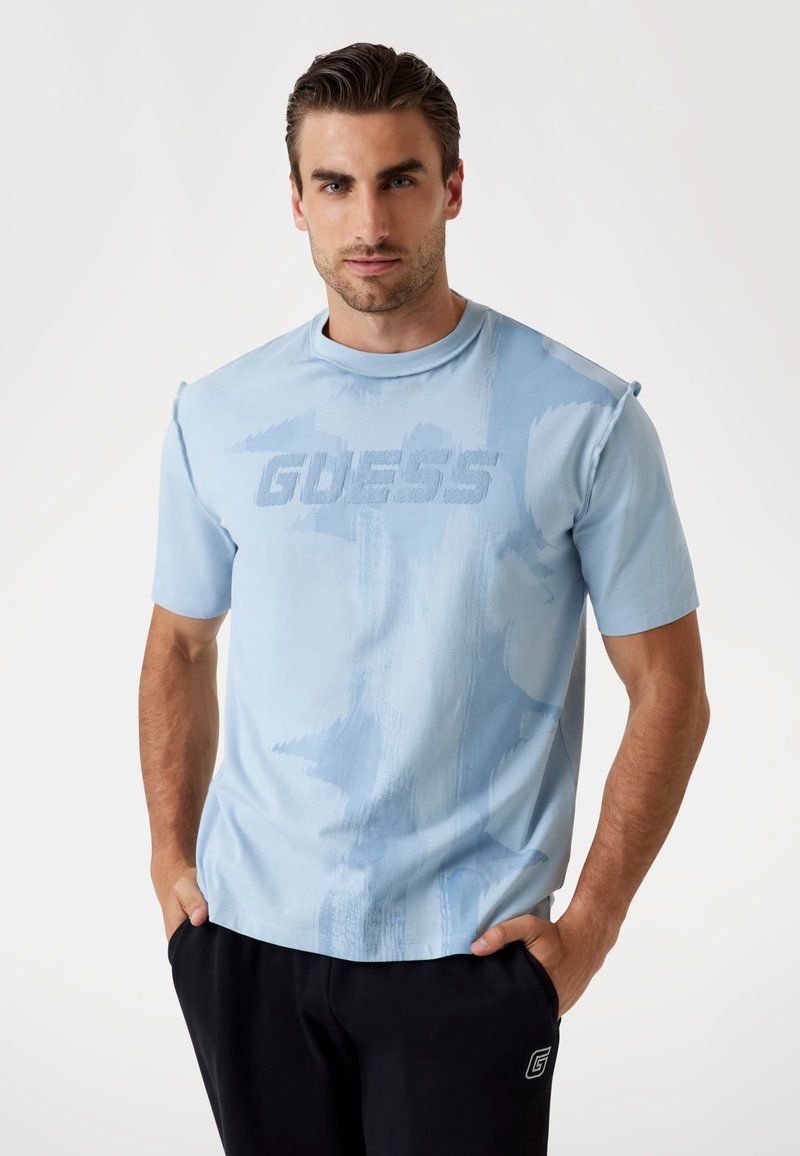 Tricou scurt cu mânecă, de culoare albastru deschis, cu o textură moale, având un logo "GUESS" tonal pe față și un model abstract subtil.