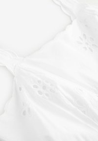 Débardeur blanc sans manches avec des découpes florales brodées décoratives, texture douce et bords en forme de scallops. Tissu lisse avec une légère brillance.