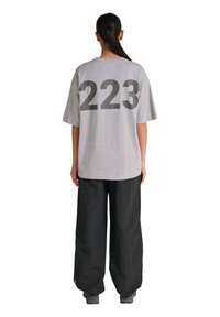 Person med sort hestehale har en oversize grå t-shirt med "223" på ryggen og løse sorte bukser, står med ryggen til.