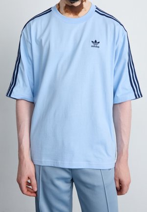 Person trägt ein hellblaues Adidas-T-Shirt mit dunkelblauen Streifen an den Ärmeln und passende hellblaue Hosen, steht vor einem schlichten Hintergrund.