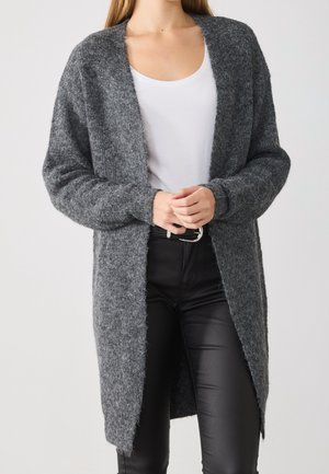 Frau trägt eine lange, offen vorn geöffnete dunkelgraue flauschige Strickjacke über einem weißen, tiefer ausgeschnittenen Top und engen schwarzen Hosen mit schwarzem Gürtel.