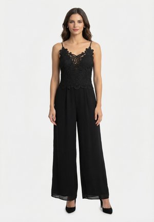 Kvinde iført sort blonderærmeløs top og løstsiddende sorte viddebenede bukser med sorte hæle, stående mod en ensfarvet hvid baggrund.