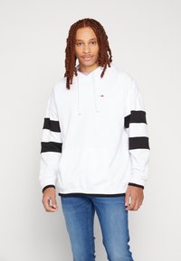 Felpa bianca con strisce nere sulle maniche, materiale di cotone, cappuccio con lacci, tasca frontale e piccolo logo sul petto. Indossata con jeans blu.