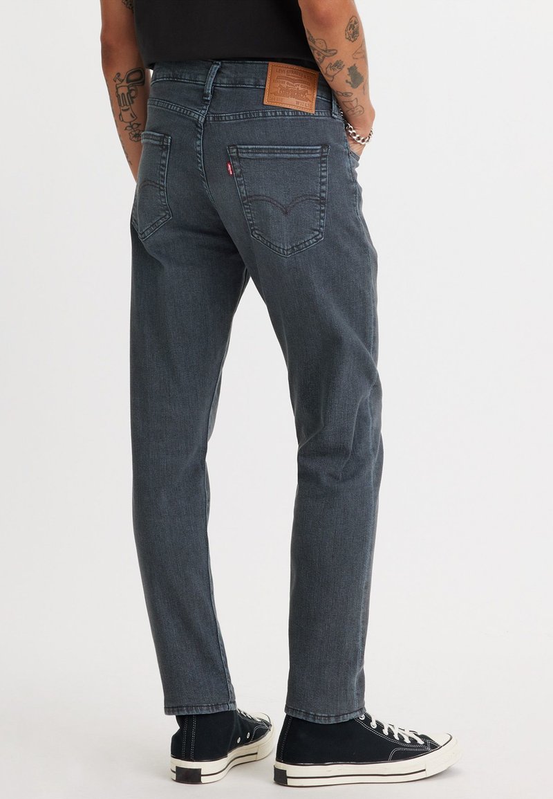 Dunkelgraue Jeans aus Denim mit geradem Schnitt, sichtbaren Nähten und Gesäßtaschen. Getragen mit schwarzen High-Top-Sneakern. Lederlabel am Bund.