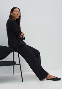 Donna in abito nero seduta di lato su una sedia nera con le braccia incrociate, davanti a uno sfondo chiaro e semplice.