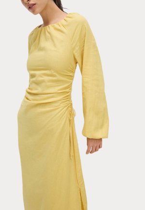 Gul hørkjole med en sammensamlet halsudskæring og lange ærmer. Har sidefrynser og en lille slids for bevægelse. Blødt, struktureret stof.