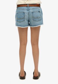 Bermudas de mezclilla con dobladillo deshilachado, color azul claro, y dos bolsillos traseros con un diseño de costura curva. Llevadas con sandalias oscuras.