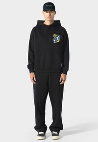 Sudadera negra con un diseño abstracto colorido en el lado izquierdo del pecho, combinada con pantalones negros de corte relajado y zapatillas negras. Textura simple y suave.