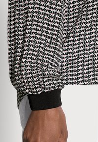 Tecido preto com um padrão floral em branco e rosa. Apresenta um punho canelado preto ao nível do pulso e um caimento ligeiramente solto com uma superfície texturada.