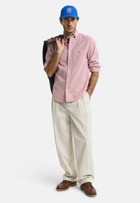 Rood-wit gestreepte lange mouwen shirt, crèmekleurige brede broek, bruine schoenen, blauwe pet met logo, marineblauwe jas over schouder.